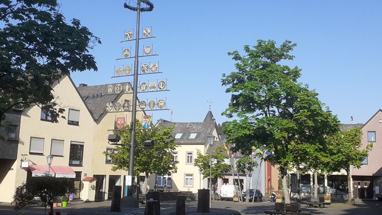 Ein Platz mit einem Maibaum, umgeben von Gebäuden und Bäumen, bei sonnigem Wetter.