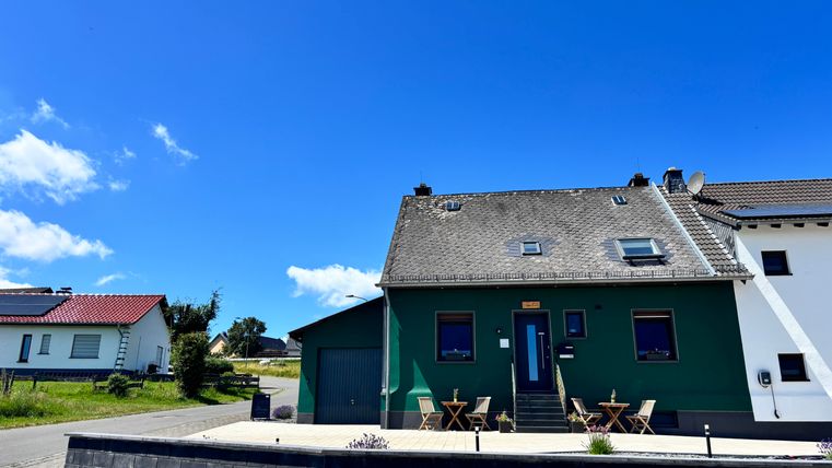 Ein grünes Haus mit einem modernen Eingangsbereich und einer Terrasse. Der Himmel ist klar und blau, umgeben von weiteren Wohnhäusern.