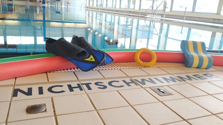 Schwimmbad mit Flossen, Poolnudeln und Schwimmbrett am Beckenrand. Auf den Fliesen steht 'NICHTSCHWIMMER'. Im Hintergrund ist das Wasser des Pools zu sehen.