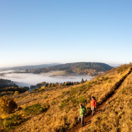 Zwei Wanderer überqueren einen hohen Vulkangipfel bei herbstlichem Wetter. Im Tal liegt ein Dorf im dichten Nebel.