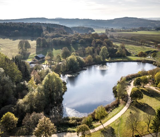 Luftaufnahme eines Sees im Bolsdorfer Tälchen, umgeben von Bäumen und Wiesen. Ein Gebäude steht am Ufer, im Hintergrund sind Hügel zu sehen., © Eifel Tourismus GmbH, Dominik Ketz