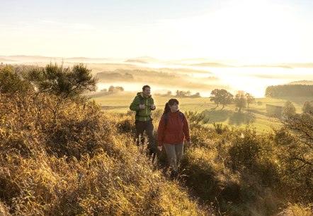 Zwei Wanderer auf einem Pfad in einer hügeligen Landschaft bei Sonnenaufgang.
