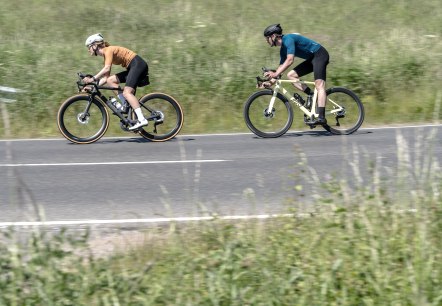 Two cyclists ride along a country road through a green, rural landscape. Fields and trees can be seen in the background., &copy; Eifel Tourismus GmbH, Dennis Stratmann