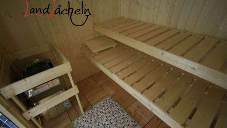 Ein heller Saunaraum mit Holzregalen und einer Sitzbank. Der Boden ist mit Fliesen ausgelegt und die Wände sind aus hellem Holz.