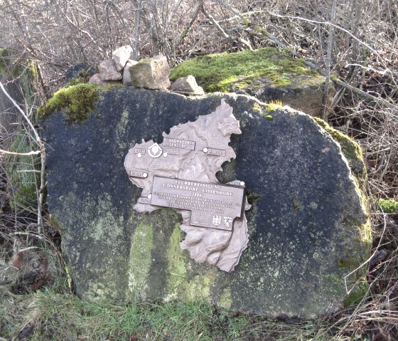 Ein Gedenkstein mit einer Reliefkarte und Inschrift, umgeben von Moos und Gras. Der Stein steht in einer natürlichen Umgebung mit Sträuchern., © Touristik GmbH Gerolsteiner Land, Ute Klinkhammer