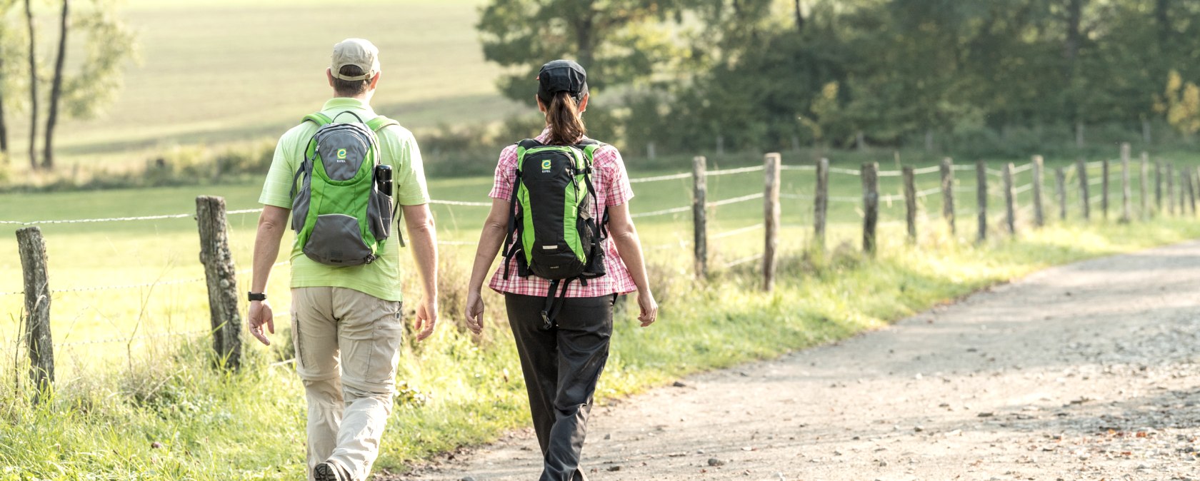 Zwei Personen wandern auf einem ländlichen Weg in der Eifel. Sie tragen Rucksäcke und sind von grüner Landschaft umgeben., © Eifel Tourismus GmbH, Dominik Ketz