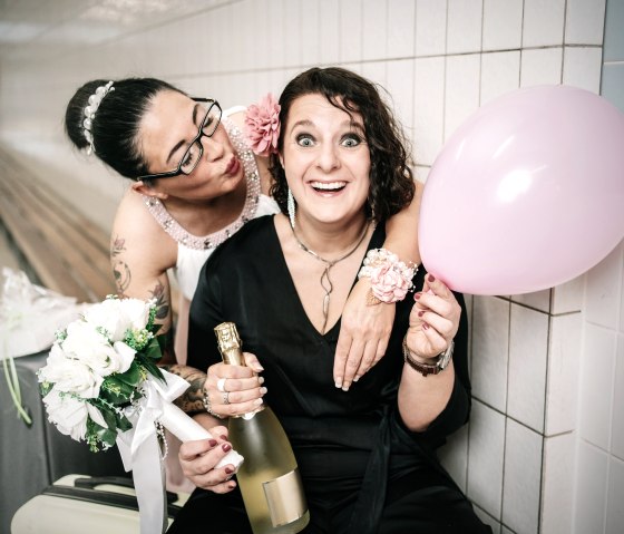Two women in stylish clothing pose together with a bottle of sparkling wine, a balloon and a bouquet of flowers in their hands.