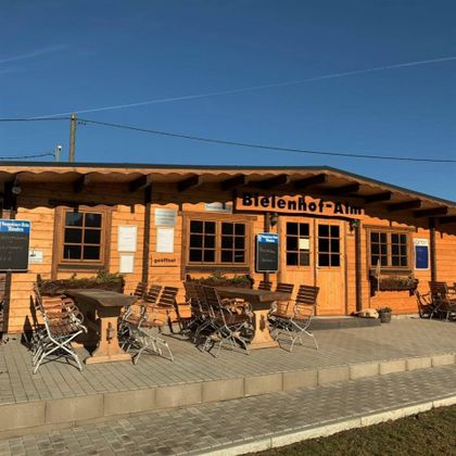 Holzgebäude der Bielenhof Alm mit Terrasse, Holztischen und Stühlen, unter blauem Himmel.