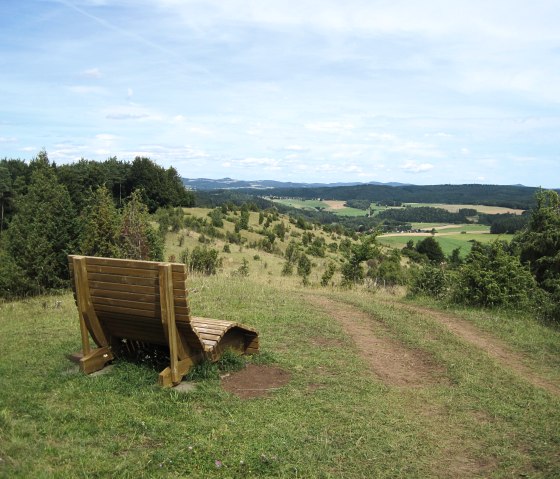 Holzbank auf einem H&uuml;gel mit Blick auf gr&uuml;ne Felder und W&auml;lder unter blauem Himmel am H&ouml;nselberg., &copy; Touristik GmbH Gerolsteiner Land, Ute Klinkhammer