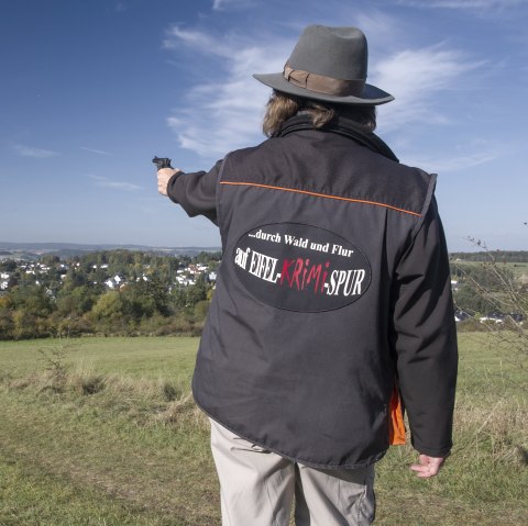Eine Hut tragende Person steht mit dem Rücken zum Betrachter und zeigt mit einer Pistole in die Ferne. Sie trägt eine Jacke mit der Aufschrift &quot;...durch Wald und Flur auf Eifel-Krimi-Spur&quot; auf dem Rücken. Vor der Person liegt eine große Wiese und dahinter in der Ferne ein Dorf mit  vielen Häusern.