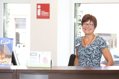 Eine Frau steht hinter einem Tresen und lächelt in die Kamera. Hinter ihr einige Fenster und das Logo der i-Marke für geprüfte Tourist-Informationen.