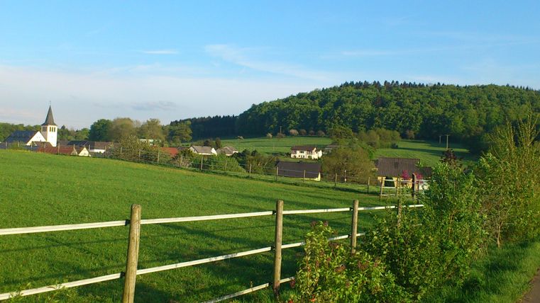 Eine idyllische Landschaft mit einem Dorf und einer Kirche im Vordergrund. Im Hintergrund sind sanfte Hügel und Bäume zu sehen.