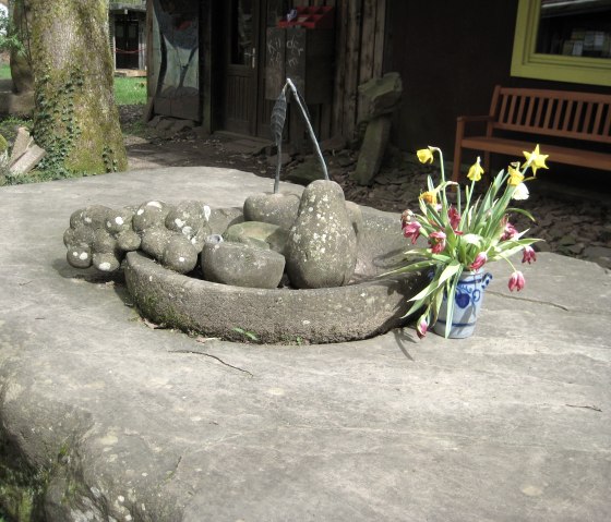Stone sculpture on a large stone, next to it a colorful bouquet of flowers in a vase. In the background, a wooden bench and a building., © Tourisik GmbH Gerolsteiner Land, Ute Klinkhammer
