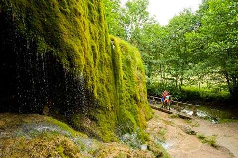 Nohner Wasserfall mit moosbewachsenen Felsen und zwei Personen auf einem Steg.