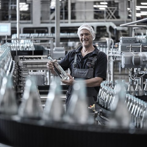 A worker in a bottling plant checks a water bottle. He is wearing work clothes and a cap. Around him are many bottles on a conveyor belt.