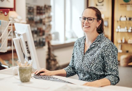 A woman stands at a high desk with a computer and keyboard in front of her and smiles at the camera. Behind her is the souvenir shop and some brochure racks from the tourist information office.
