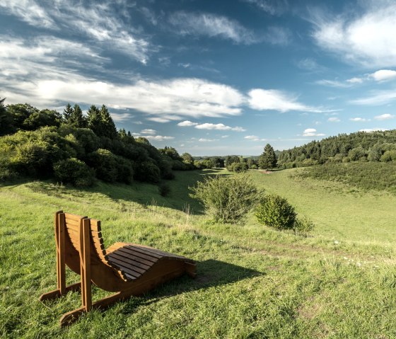 Holzliege auf einer Wiese mit Blick auf die gr&uuml;ne Papenkaule und einen blauen Himmel mit Wolken., &copy; Eifel Tourismus GmbH, Dominik Ketz