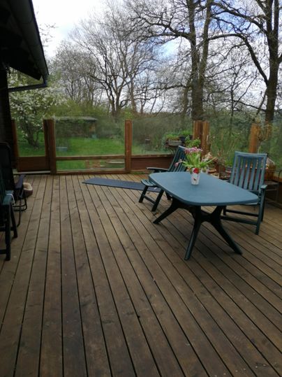 Een gezellige houten terras met een tafel en stoelen. Op de achtergrond zijn bomen en een groene wei zichtbaar.