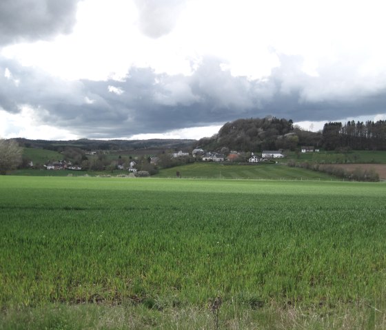 Green meadows stretch up to a wooded hill. Houses and dark clouds can be seen on the horizon., © Touristik GmbH Gerolsteiner Land, Ute Klinkhammer