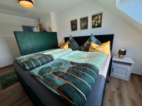 Ein gemütliches Schlafzimmer mit einem Doppelbett und dekorativen Kissen. An der Wand hängen Bilder, und es gibt einen Nachttisch mit einer Lampe.