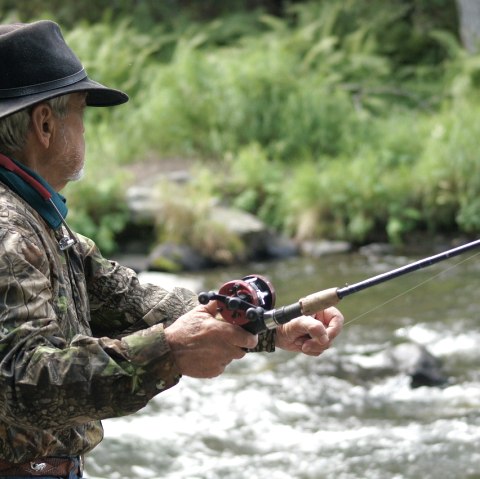 A man in camouflage clothing and a fishing hat stands by or in a river and casts his line.