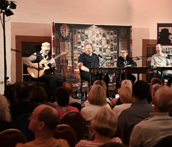 Drei Männer und eine Frau sitzen nebeneinander an Tischen auf einer Bühne vor dichtem Publikum. Der Mann links spielt Guitarre und die anderen schauen im zu.