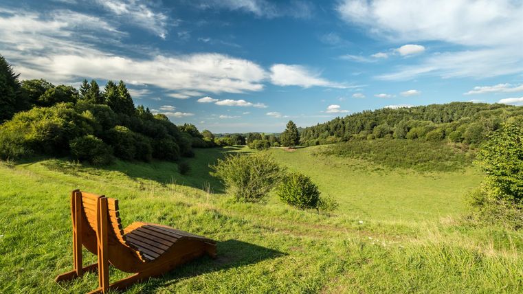 Chaise longue en bois au bord du cratère volcanique vert de Papenkaule, entourée d'arbres et de prairies sous un ciel bleu avec des nuages.