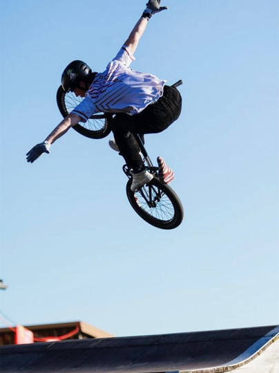 Ein BMX-Fahrer zeigt einen spektakulären Trick in großer Höhe über einer Rampe. Mit ausgestreckten Armen und kontrollierter Körperhaltung demonstriert er Geschick, Mut und Präzision vor klarem blauem Himmel.