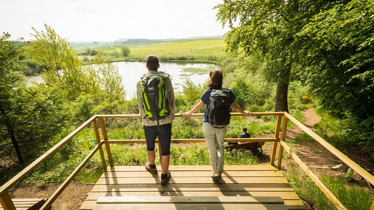 Im vordergrund stehen eine Frau und ein Mann auf einer hölzernen Plattform. Im Hintergrund ist eine runde Wasserfläche zu sehen.Am rechten und linken Bildrand stehen einige Bäume. Es ist Frühling, der Ginster blüht. Hinter dem Maar liegen Wiesen und der Blick reicht weit in das Gerolsteiner Land.