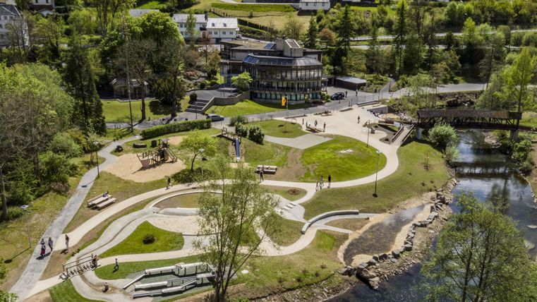 Luftaufnahme eines Kurparks mit geschwungenen Wegen, Spielplatz, Fluss und umliegenden Gebäuden. Menschen spazieren und genießen die Natur.