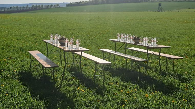 Een gedekte tafel op een weiland met drankjes en houten banken. Op de achtergrond zijn bossen en een uitgestrekt landschap te zien.