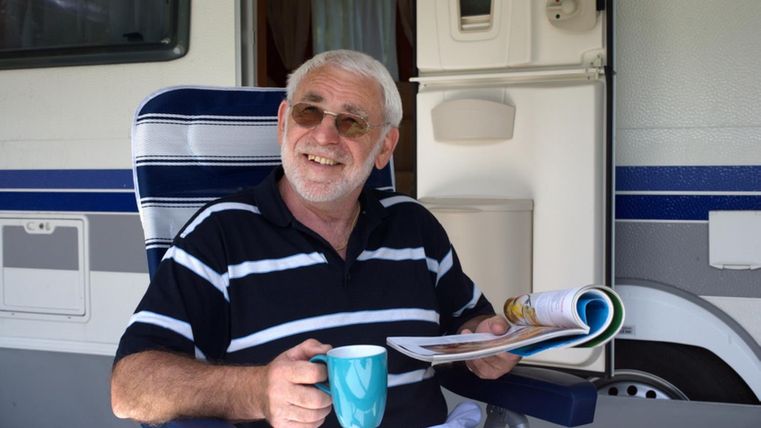 Ein Mann sitzt entspannt vor einem Wohnmobil, hält eine Tasse und ein Magazin. Er trägt eine Sonnenbrille und lächelt in die Kamera.