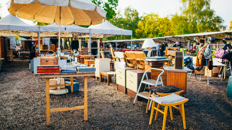Een vlooienmarkt met verschillende meubels en stoelen onder parasols. Op de achtergrond zijn andere kramen en bezoekers te zien.