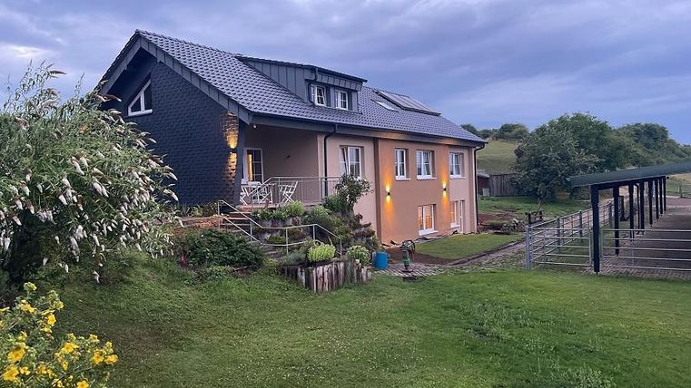 Ein schönes Haus mit einem gepflegten Garten. Im Hintergrund sind hügelige Wiesen und ein Stall zu sehen.