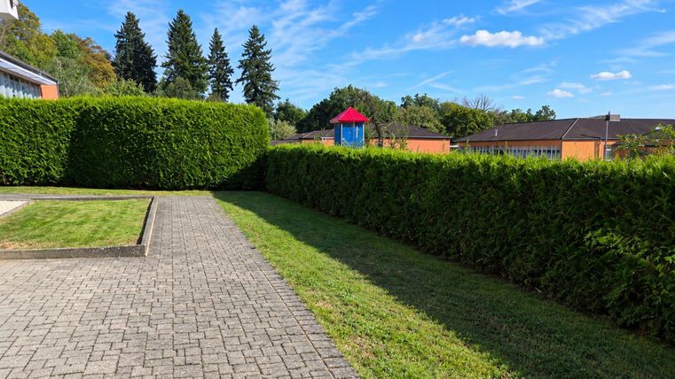 Une surface pavée avec une pelouse verte et des haies hautes. En arrière-plan, on peut voir des bâtiments et un terrain de jeu.