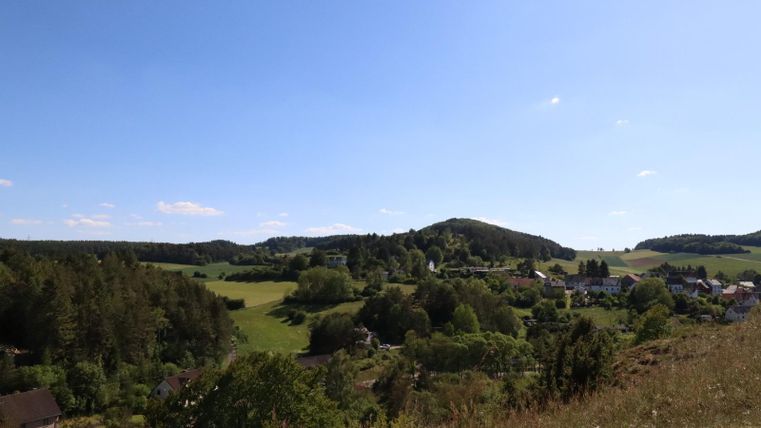 Eine malerische Landschaft mit sanften Hügeln und grünem Wald. Im Vordergrund sind kleine Häuser und weite Wiesen zu sehen.