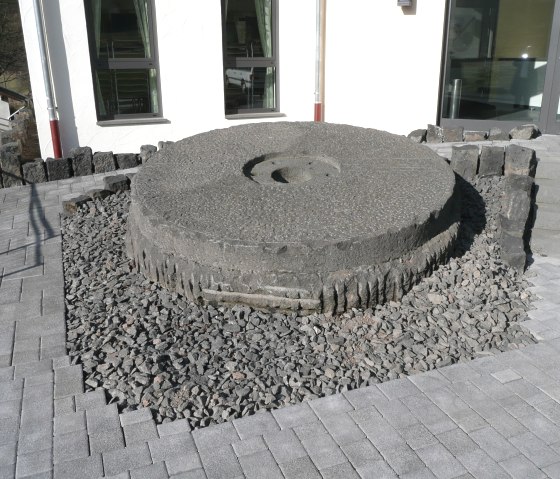 Een grote, ronde molensteen ligt op een grindbed voor een modern gebouw met ramen en een deur., © Touristik GmbH Gerolsteiner Land