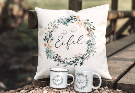 Handmade fabric cushion with floral pattern and &lsquo;Eifel&rsquo; lettering in the centre on a wooden bridge. In front of it are two mugs with the same design from Mitgebracht.