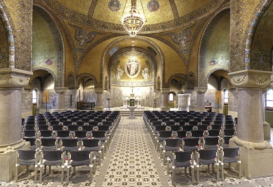 Das Bild zeigt das Innere der Erlöserkirche mit Blick zur Decke und zum Altar. Die Decke ist mit goldenen Mosaiken, Engeln und einem großen Kronleuchter geschmückt. Fenster lassen Licht in den prunkvollen Kirchenraum. Zahlreiche Stühle bilden Sitzreihen für Besucher.