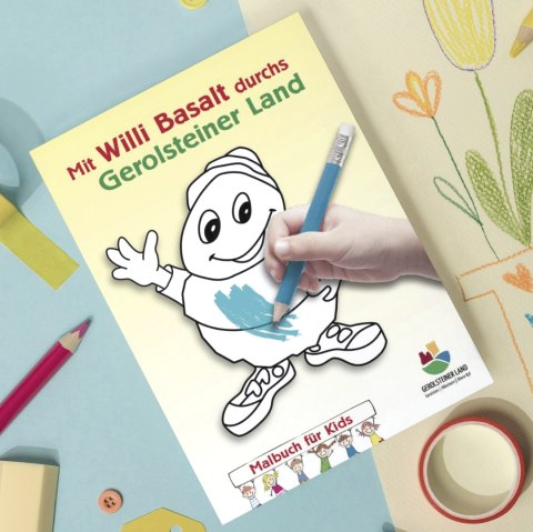 Voorpagina van het kleurboek “Met Willi Basalt door het Gerolsteiner Land” op een tafel vol kleurspullen en kleurrijke vellen papier.