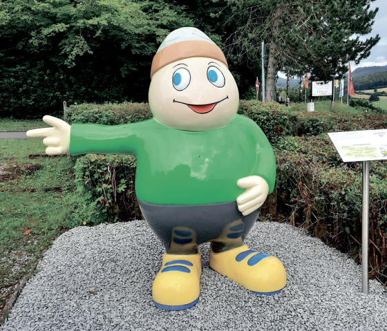 Statue von Maskottchen Willi Basalt mit gr&uuml;nem Shirt, grauer Hose und gelben Schuhen zeigt nach links. Hinter ihm B&auml;ume und ein Parkplatz.