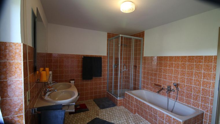 Ein modernes Badezimmer mit zwei Waschbecken, einer Badewanne und einer Duschkabine. Die Wände sind mit orangefarbenen Fliesen gefliest, und der Boden ist mit hellen Steinfliesen gestaltet.