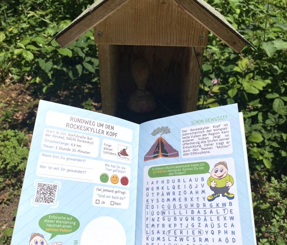 An open hiking pass with information about the Rockeskyller Kopf circular trail and a search puzzle, held in front of a small wooden house in a green setting., © Touristik GmbH Gerolsteiner Land, Leonie Post