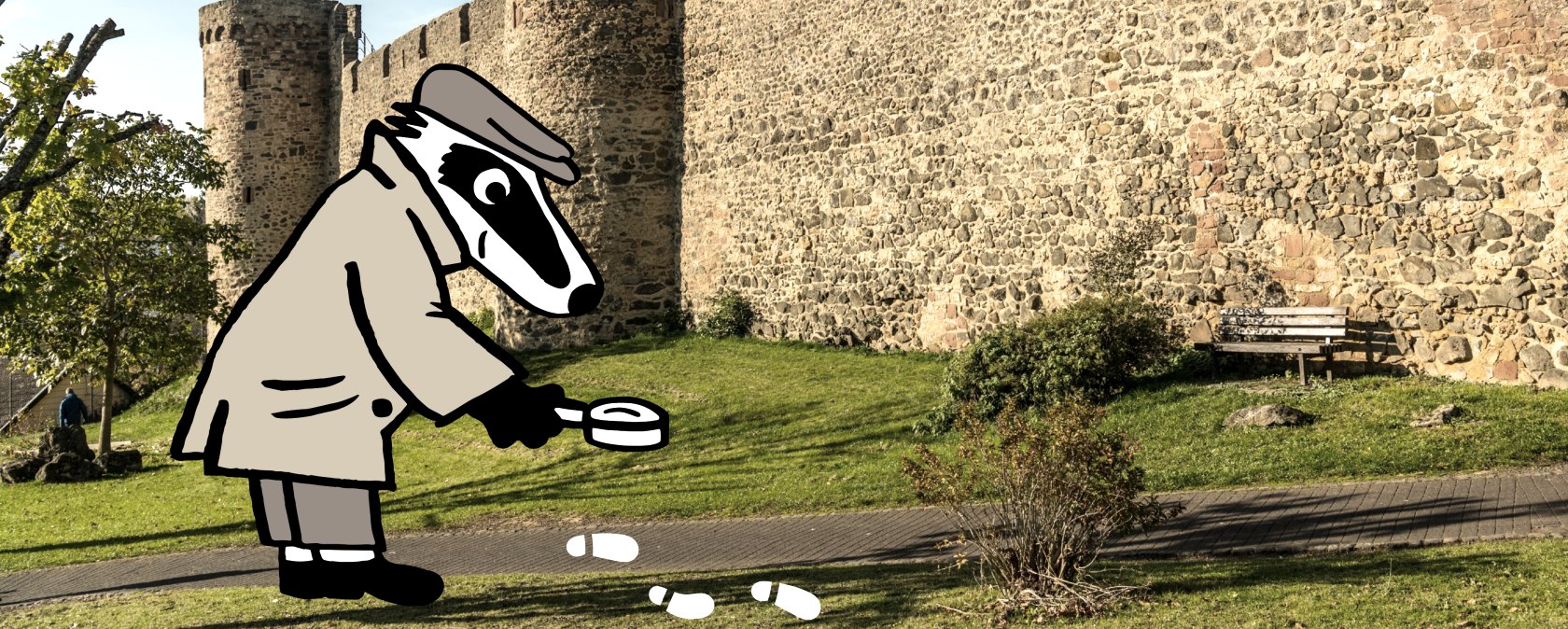 De hoge stadsmuur van Hillesheim staat voor een groen gebied onder een blauwe hemel. Rechercheur Badger onderzoekt met een vergrootglas witte voetafdrukken op een smal pad voor de muur.