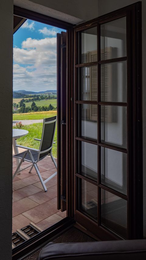 Eine offene Tür, die auf eine Terrasse mit einem Tisch und einem Stuhl blickt. Im Hintergrund sind grüne Felder und ein blauer Himmel zu sehen.