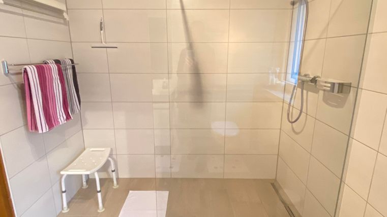 Ein modernes Badezimmer mit einer großen Dusche und hellen Fliesen. Zwei Handtücher hängen an der Wand, und ein Hocker steht in der Dusche.