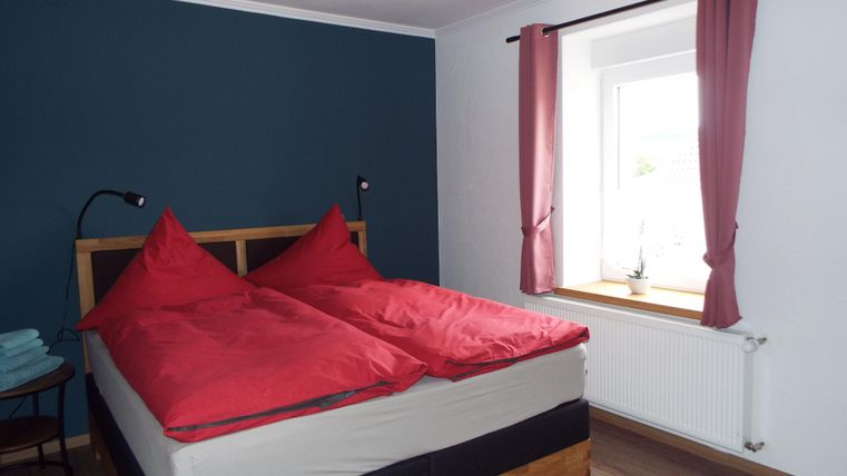 Ein gemütliches Schlafzimmer mit einem großen Bett und roten Kissen. Das Zimmer hat blauen Wände und einen hellen Fensterbereich.