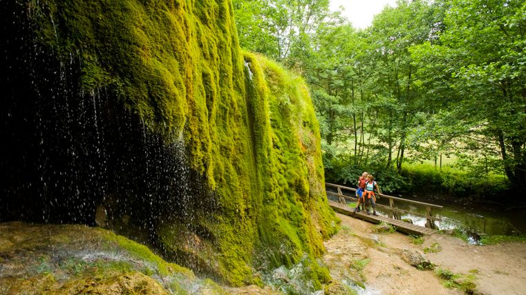 Nohner Wasserfall mit moosbewachsenen Felsen und zwei Personen auf einem Steg.
