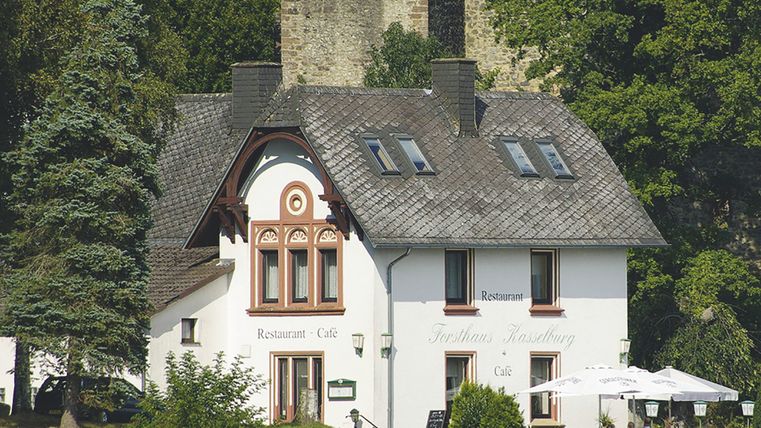 <p>Im Vordergrund sihet man ein weißes Haus mit der Aufschrift "Restaurant Café" und "Forsthaus Kasselburg", Davor stehen zwei große Schirme und Tische mit Stühlen, Im Hintergrund ist eine Burg und Bäume</p>