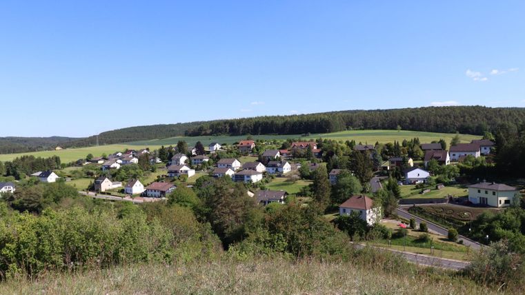 Eine malerische Landschaft mit einem kleinen Dorf, umgeben von grünen Feldern und Wäldern. Der Himmel ist klar und blau.